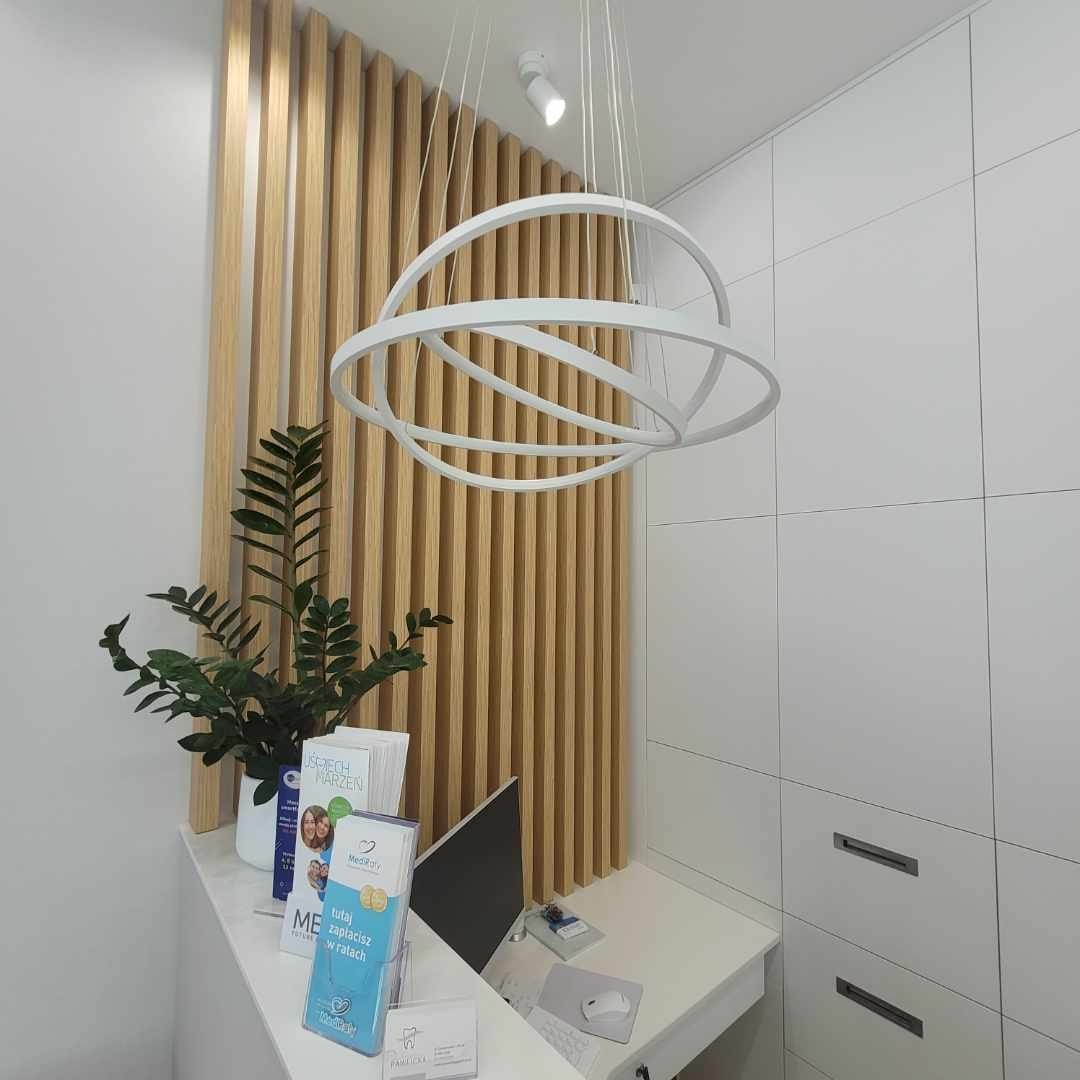 Recepcja w jasnej tonacji z drewnianymi panelami, rośliną doniczkową i nowoczesną lampą pierścieniową, ulotki informacyjne na blacie.