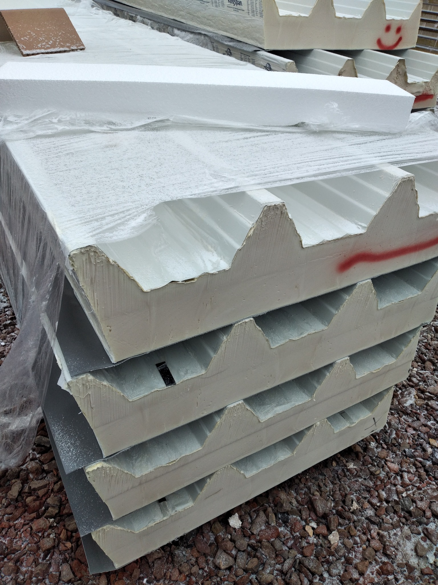 Stos płyt warstwowych z widocznym przekrojem, zabezpieczonych folią, z czerwoną, uśmiechniętą buźką narysowaną na jednej z nich, ułożonych na żwirowym podłożu.