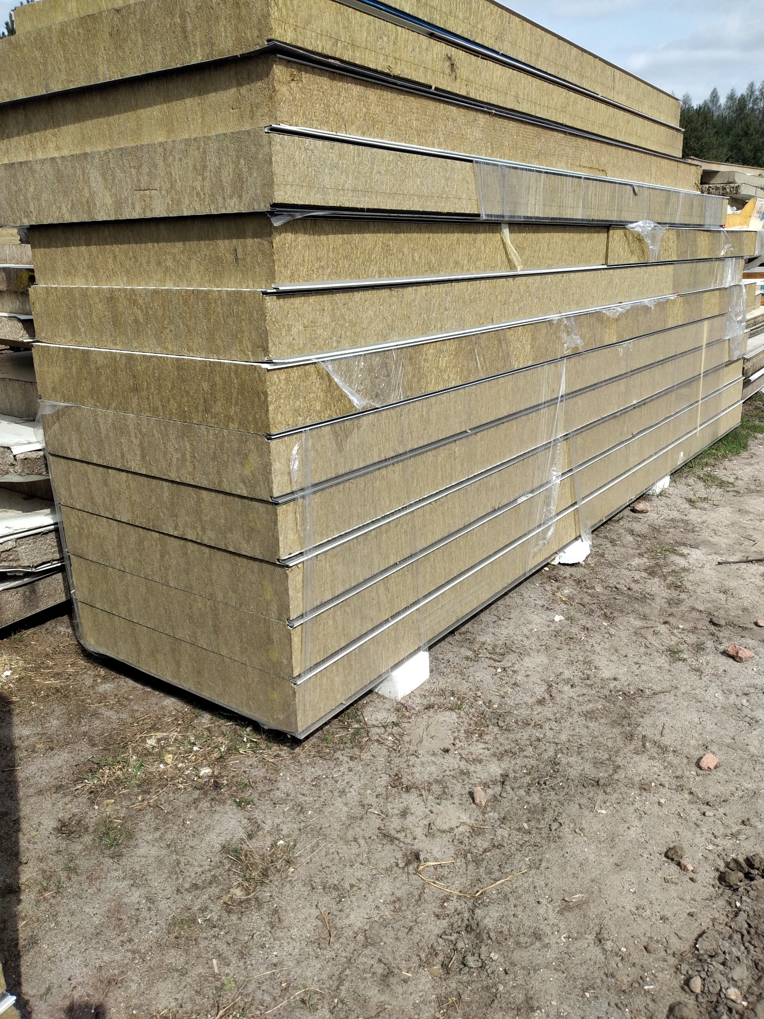 Stos płyt warstwowych z wełny mineralnej, zabezpieczonych folią, oczekujących na transport w Mierzęcicach, widoczny fragment gruntu.