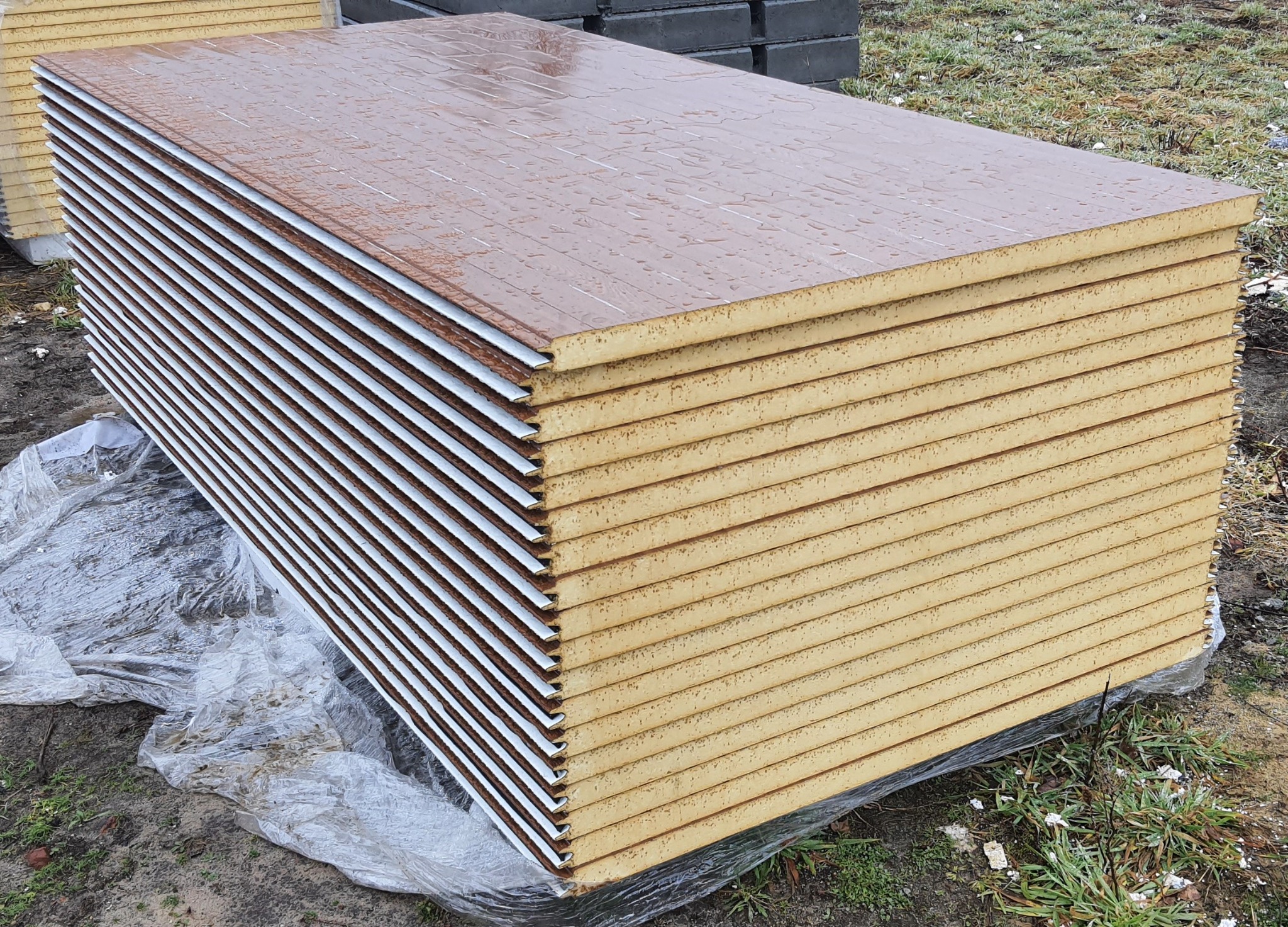 Stos brązowych płyt warstwowych z widocznym rdzeniem izolacyjnym, zabezpieczonych folią na zewnątrz, leżący na trawie.
