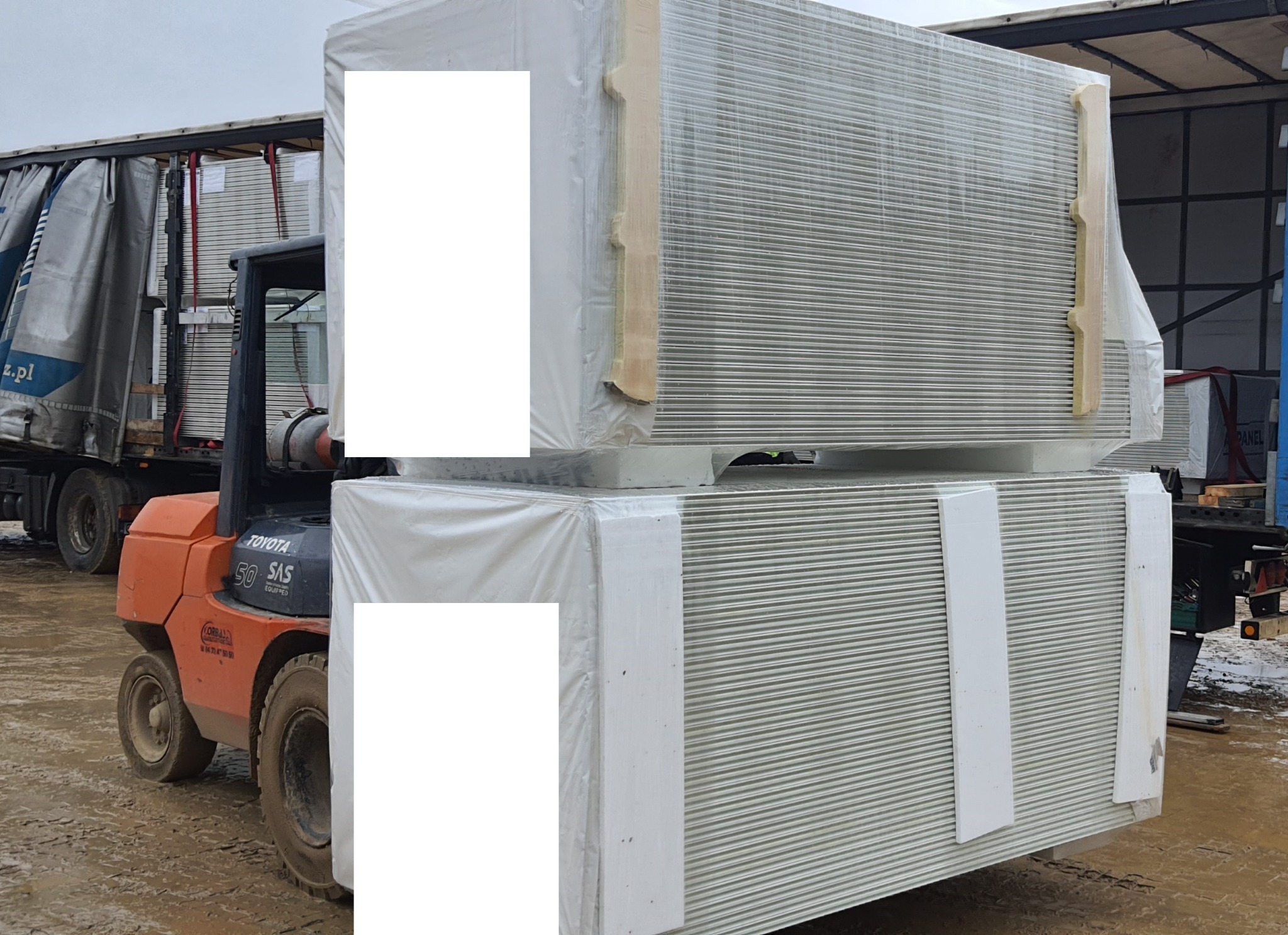 Wózek widłowy Toyota 50 SAS transportuje dwa zabezpieczone folią i drewnianymi listwami pakiety blachy trapezowej na terenie składu budowlanego.