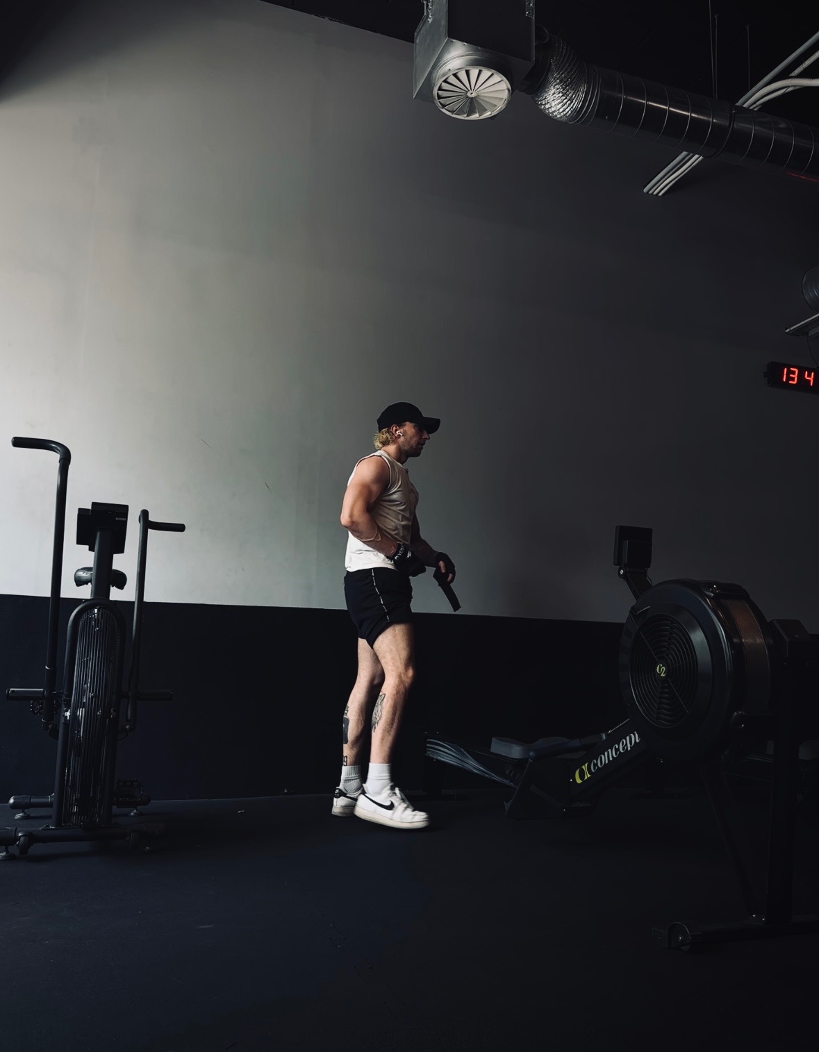 Mężczyzna w tank topie i czapce bejsbolowej stoi w siłowni obok ergometru wioślarskiego Concept2, z tatuażami na nogach i zegarem cyfrowym w tle.