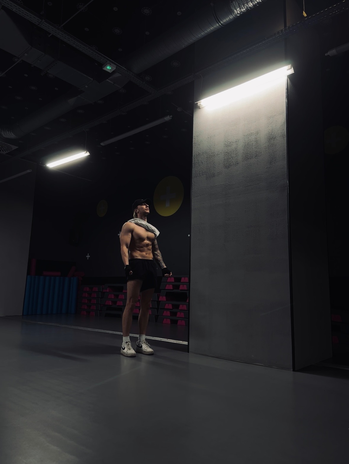Mężczyzna z wytatuowaną ręką, bez koszulki, w czapce i rękawiczkach, stoi w siłowni pod lampą, z ręcznikiem na szyi, patrząc w górę.