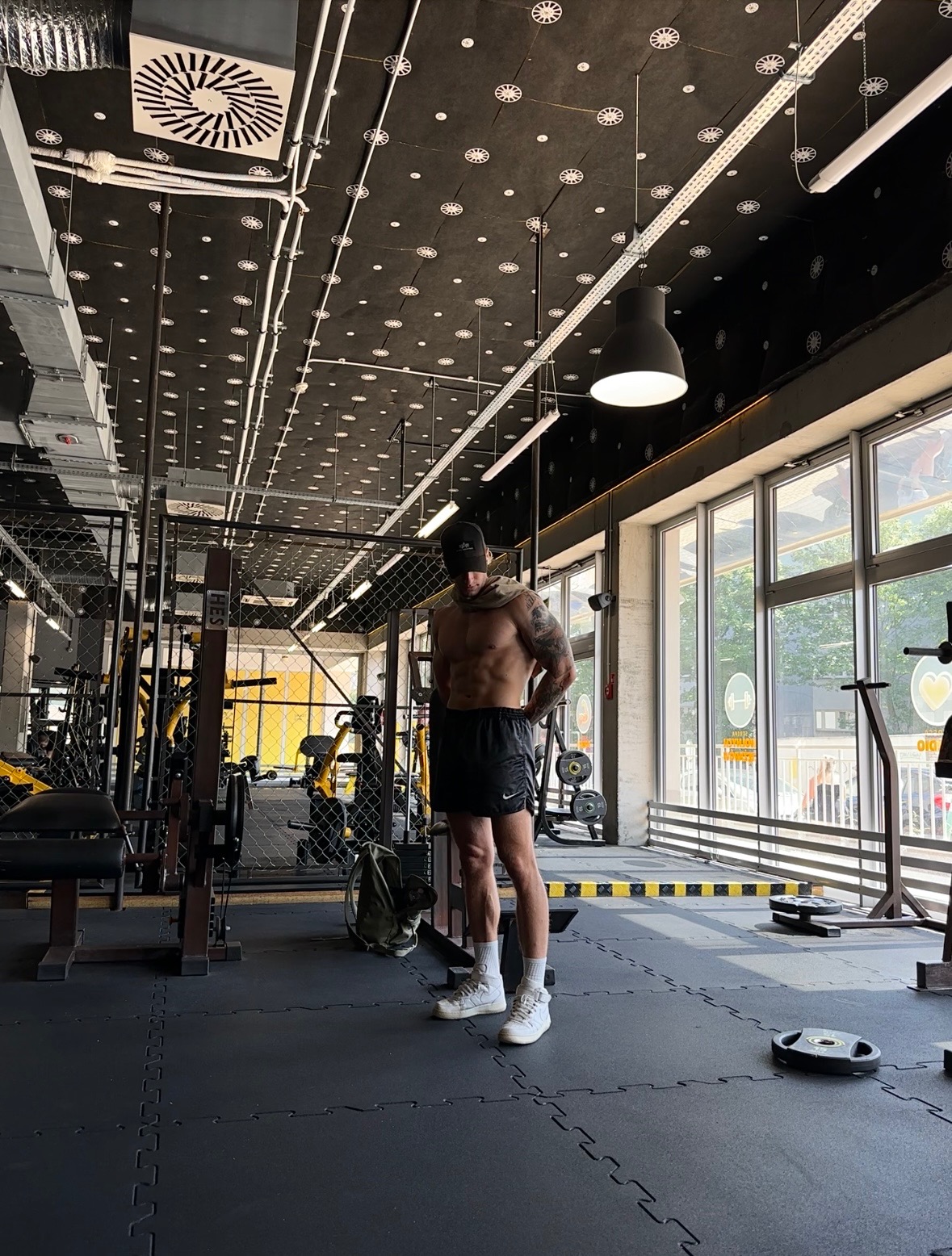 Mężczyzna w sportowych szortach i czapce stoi w siłowni na tle sprzętu do ćwiczeń, z widocznymi tatuażami na ramionach i nietypowym wzorem na suficie.