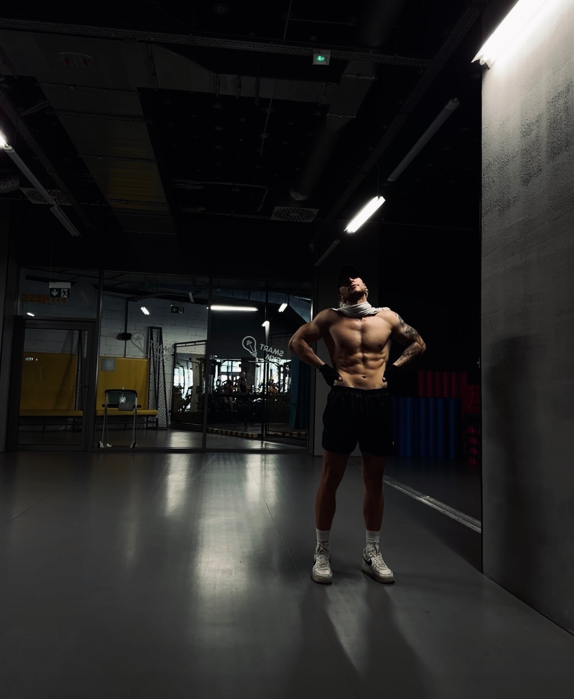 Mężczyzna z odsłoniętym torsem, w czapce i rękawiczkach, stoi w siłowni z rękami na biodrach, patrząc w górę.