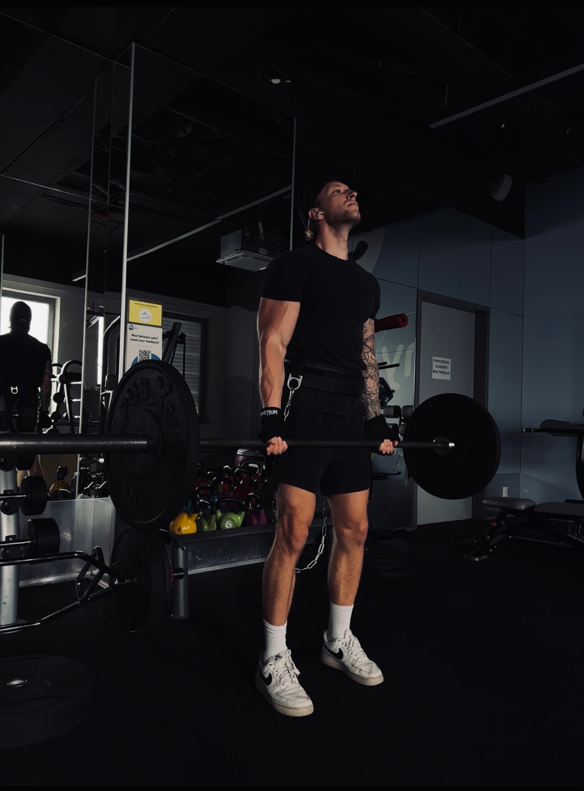 Mężczyzna w czarnym stroju, z tatuażem na ramieniu, podnosi sztangę w ciemnej siłowni, z kettlebells w tle i lustrzanym odbiciem drugiej osoby.