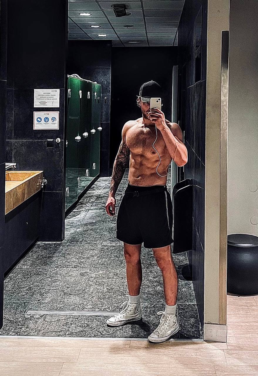 Mężczyzna bez koszulki w czapce robi selfie w lustrze na siłowni, widoczne tatuaże na ramieniu i słuchawki.