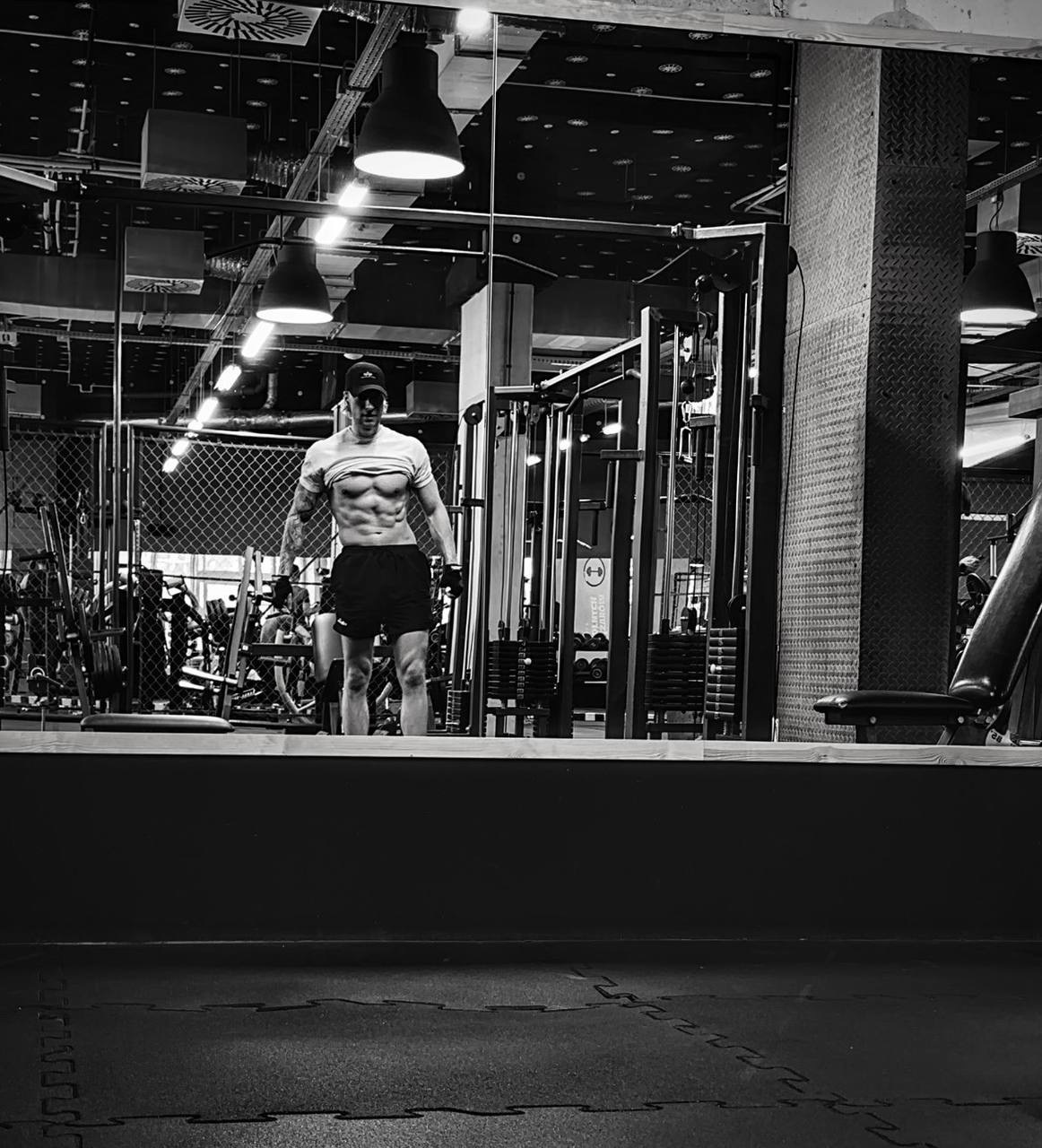 Mężczyzna z wyraźnie zarysowanymi mięśniami brzucha, pozujący w lustrze siłowni, ubrany w czapkę z daszkiem i rękawiczki treningowe, widoczne maszyny do ćwiczeń w tle.