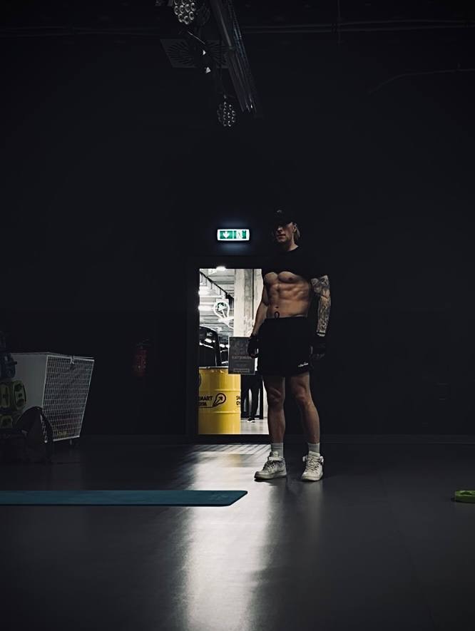 Mężczyzna z widocznymi mięśniami brzucha, w czarnej koszulce i spodenkach, stoi w ciemnym pomieszczeniu siłowni, na podłodze mata do ćwiczeń, w tle żółta beczka z napisem.