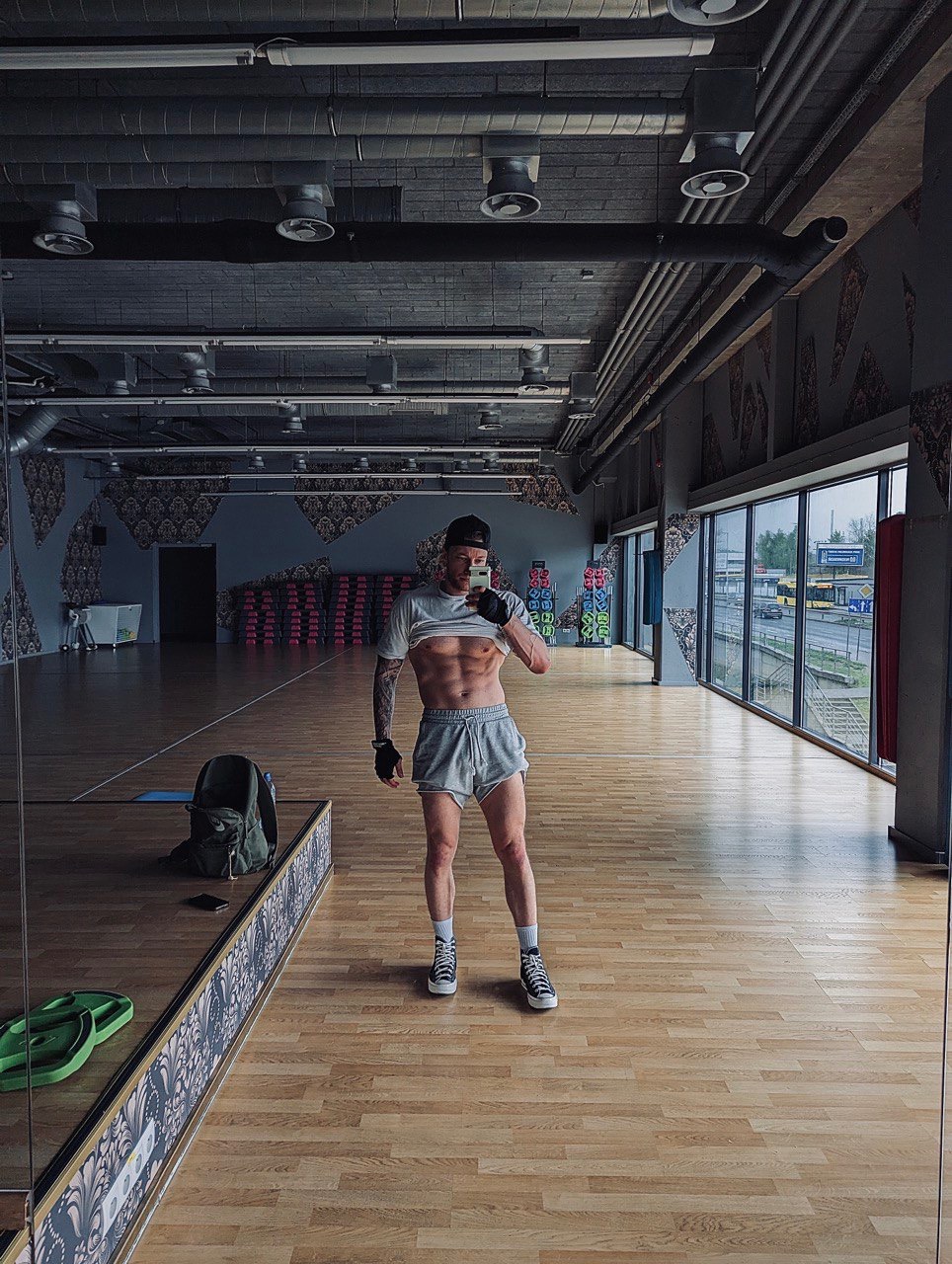 Mężczyzna w sportowym stroju, z tatuażami na ramieniu, pozuje w przestronnej siłowni, unosząc koszulkę, by pokazać wyrzeźbiony brzuch, robiąc selfie w lustrze.