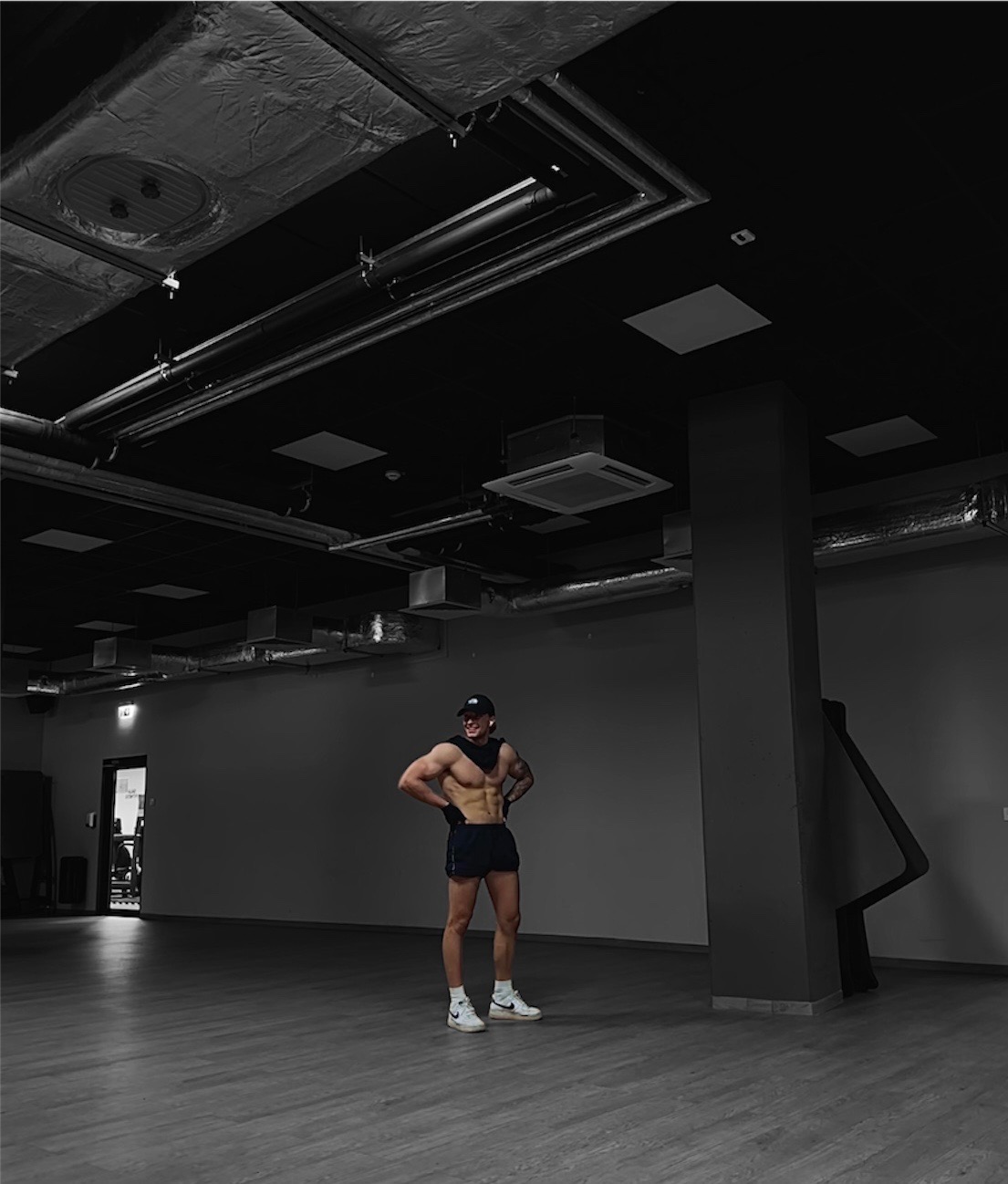 Mężczyzna z widoczną muskulaturą pozuje w pustej siłowni, widoczne instalacje pod sufitem, czarno-białe zdjęcie.