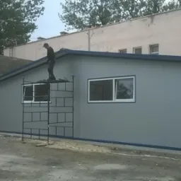 Montaż hali z płyt warstwowych: pracownik na rusztowaniu przy oknie, szara elewacja z niebieskimi akcentami, widok z zewnątrz, teren budowy.