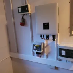 Biała ściana z zamontowanymi urządzeniami elektrycznymi: inwerter fotowoltaiczny FoxESS, rozdzielnice elektryczne z oznaczeniami PV, przewody w białych rurkach PCV.