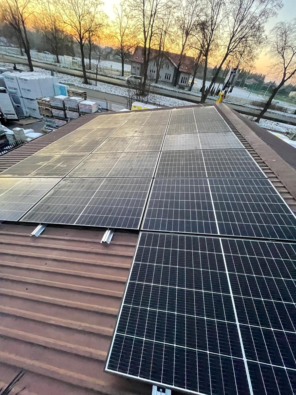 Panele fotowoltaiczne zamontowane na brązowym dachu z blachy trapezowej, widoczne elementy montażowe, w tle budynki i drzewa oświetlone wschodzącym słońcem.
