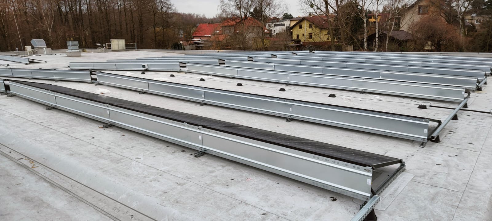 Montaż konstrukcji wsporczej pod panele słoneczne na płaskim dachu budynku przemysłowego; widoczne szyny montażowe i elementy mocujące, tło stanowią budynki mieszkalne i drzewa bez liści.