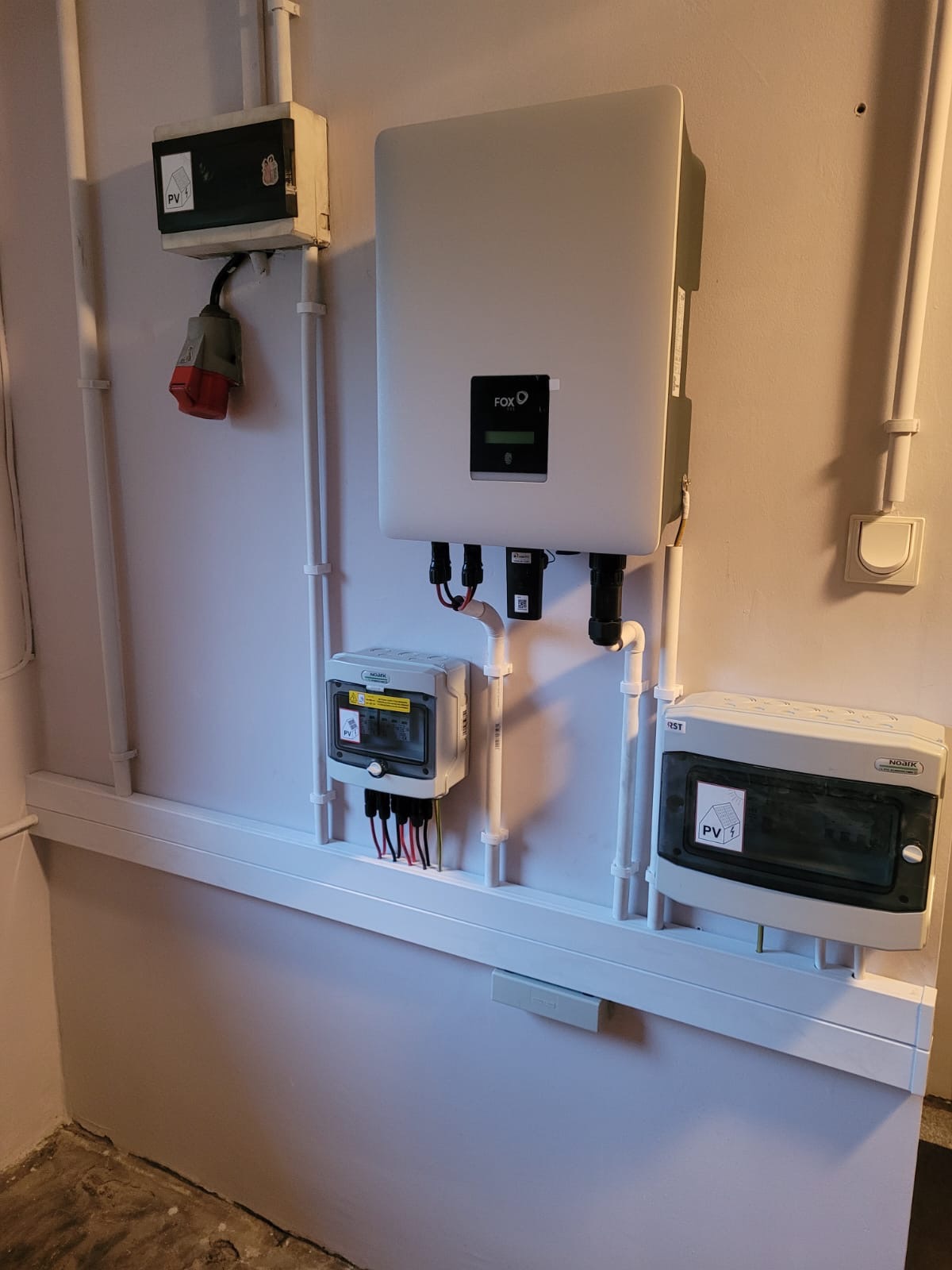 Szary falownik FOX podłączony do instalacji elektrycznej wewnątrz budynku, obok widoczne liczniki i zabezpieczenia w białych obudowach z naklejkami PV.