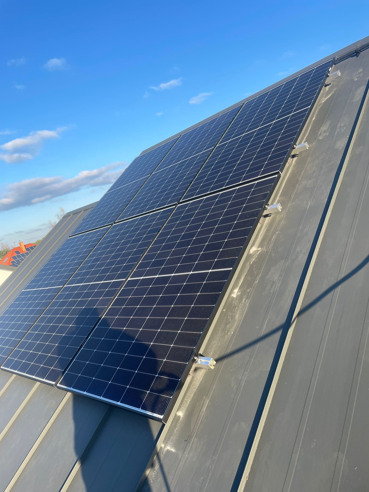 Panel słoneczny zamontowany na szarym metalowym dachu pod błękitnym niebem, widoczne mocowania i cień. W tle fragment dachu innego budynku.