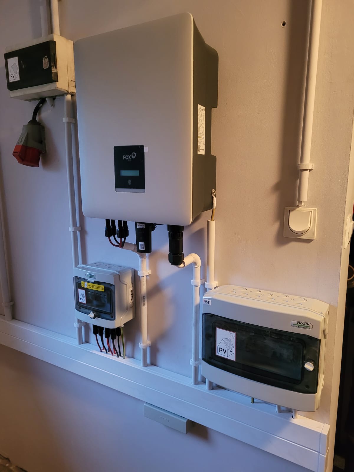 Szara ściana z zamontowanym inwerterem marki Fox, dwoma skrzynkami zabezpieczeń z naklejkami PV oraz okablowaniem w białych listwach.