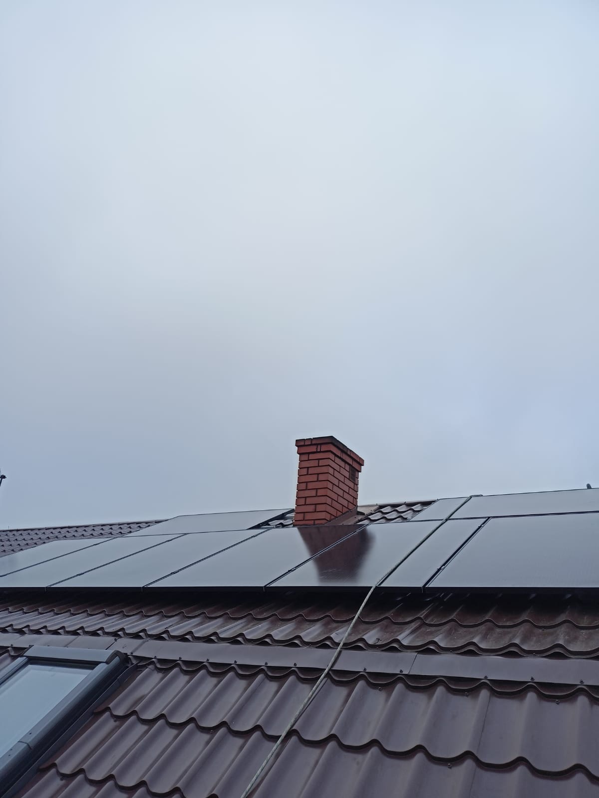 Montaż paneli fotowoltaicznych na dachu z brązowej blachodachówki, widoczny komin z czerwonej cegły i okno dachowe, pochmurne niebo.