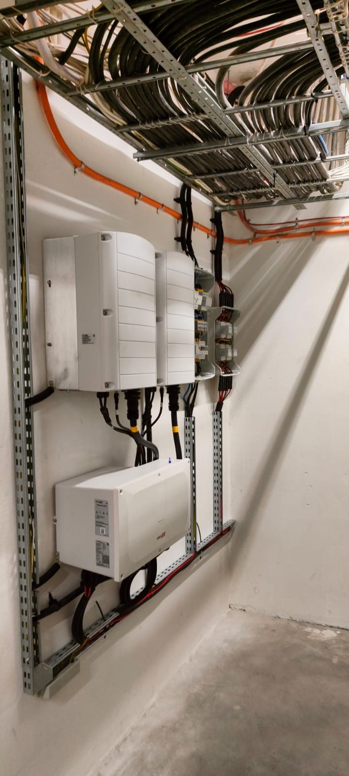 Wewnętrzna instalacja z inwerterami na ścianie, widoczne kable w korytach i przewody elektryczne, surowy beton na podłodze.