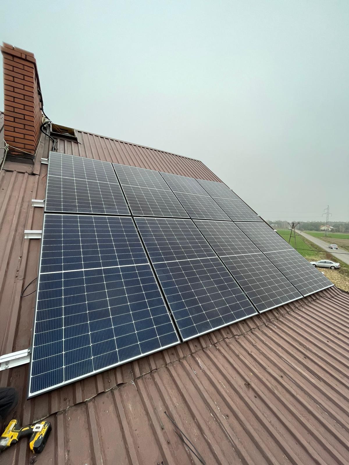 Panele słoneczne zamontowane na brązowym, blaszanym dachu domu jednorodzinnego z ceglanym kominem w pochmurny dzień. W tle widoczna droga i słup energetyczny.