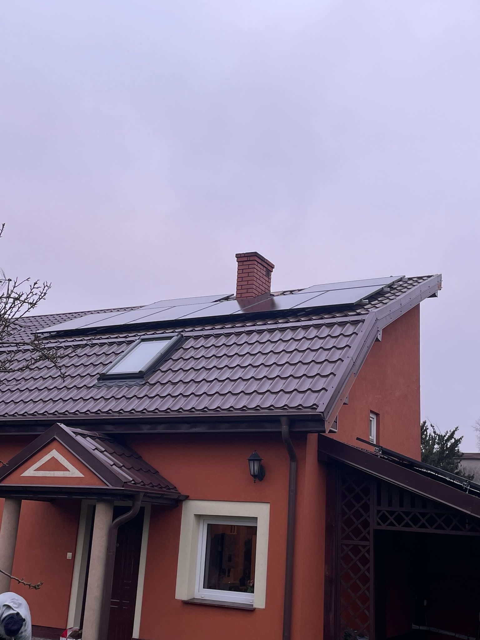 Czerwony dom z brązowym dachem, na którym zamontowano panele słoneczne i okno dachowe, widok z dołu w pochmurny dzień.