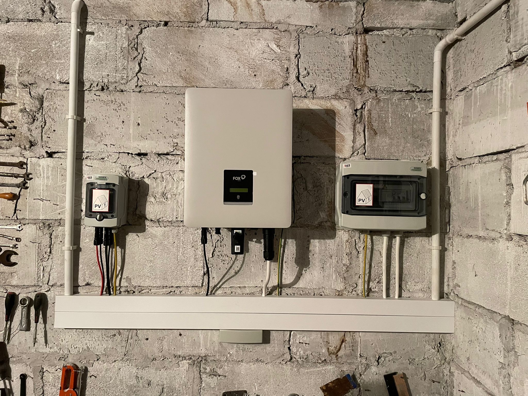 Szary inwerter marki FOX i dwa małe rozdzielacze z oznaczeniem PV zamontowane na szorstkiej, pomalowanej na biało ścianie z cegły. Widoczne przewody elektryczne i narzędzia na ścianie.