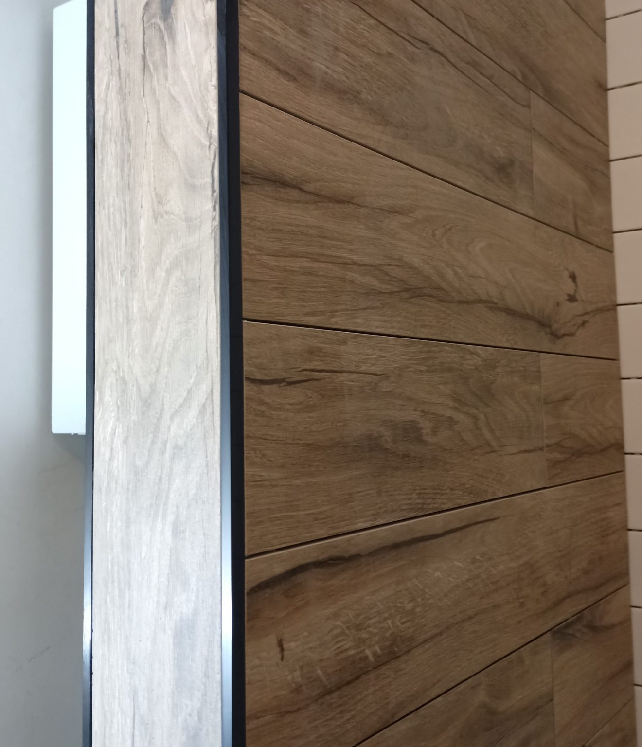 Ściana wyłożona płytkami imitującymi drewno w odcieniach brązu, ułożonymi poziomo, z widoczną białą szafką i fragmentem ściany z białymi płytkami w tle.