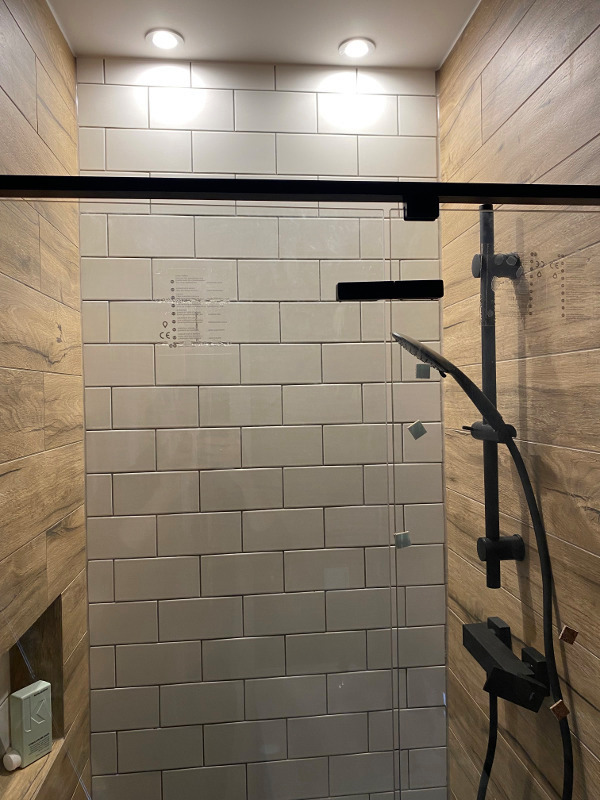 Kabina prysznicowa z czarnym osprzętem, białe prostokątne płytki na ścianie, drewnopodobne panele po bokach, widoczna butelka kosmetyku w niszy.