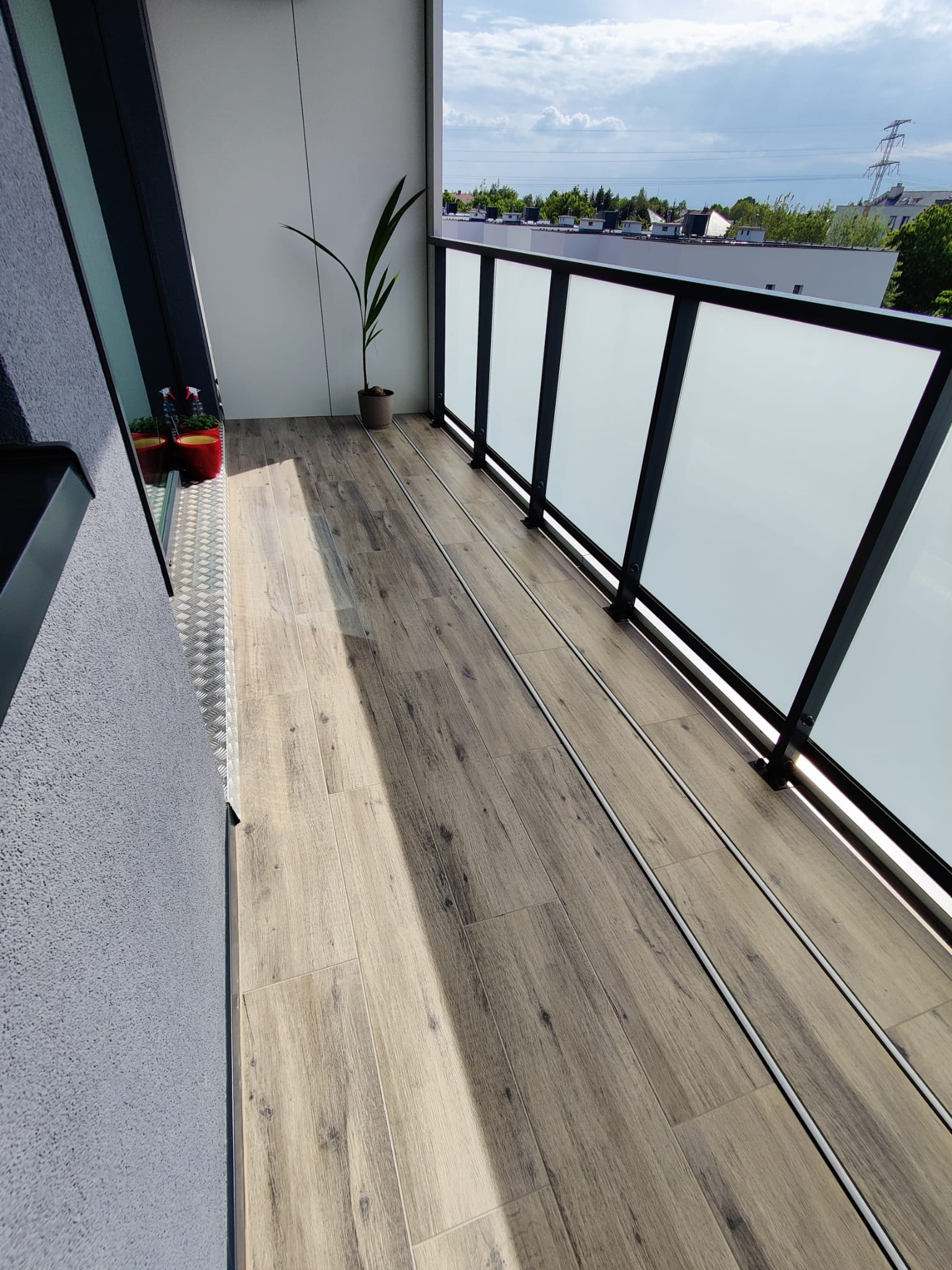 Balkon z podłogą wyłożoną płytkami imitującymi drewno, szklaną balustradą z czarną ramą oraz roślinami w doniczkach przy ścianie.