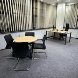 Jasne biuro z szarym dywanem, okrągłym stołem konferencyjnym z czterema czarnymi krzesłami, biurkiem z komputerem i roletami pionowymi.