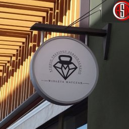 AUKCJE I SOCIAL MEDIA KAROL LASER - Okrągły szyld z logo studia treningu personalnego 'Wioleta Matczak' zamontowany na elewacji budynku z drewnianą okładziną.