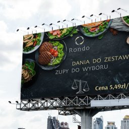 AUKCJE I SOCIAL MEDIA KAROL LASER - Billboard reklamowy z ofertą dań i zup restauracji Rondo na tle miejskiego krajobrazu i pochmurnego nieba.