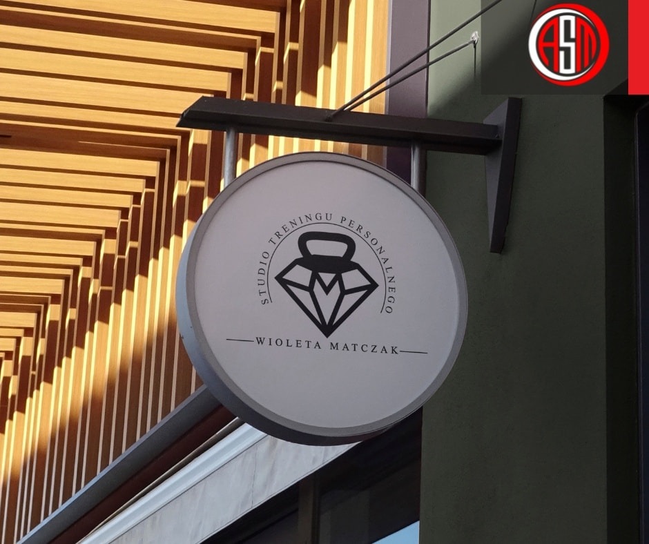 Okrągły szyld z logo studia treningu personalnego 'Wioleta Matczak' zamontowany na elewacji budynku z drewnianą okładziną.