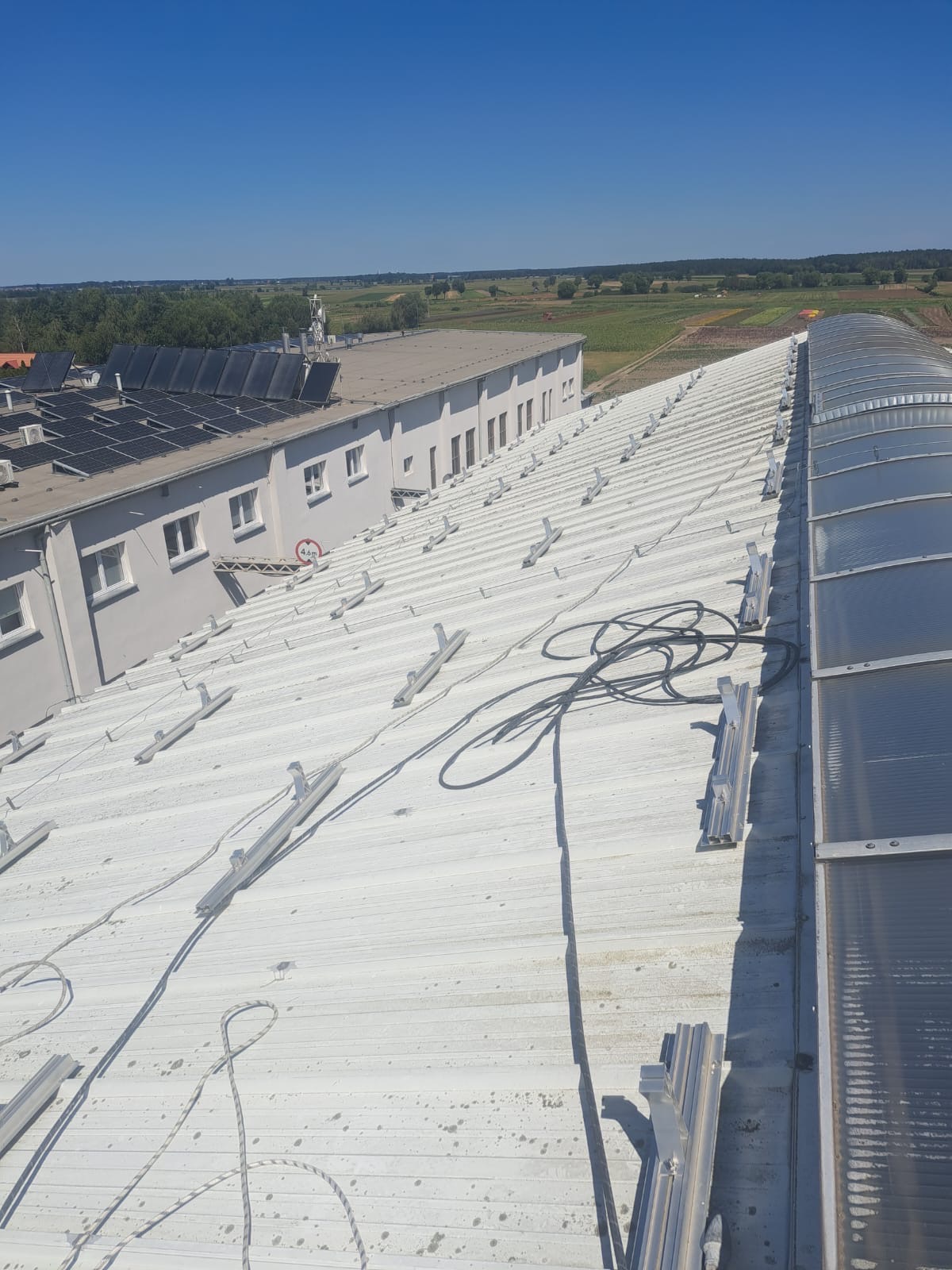 Metalowy dach przemysłowy z zamontowanymi uchwytami pod instalację, widoczne przewody i fragment przeszklonej konstrukcji dachowej, w tle budynek z panelami słonecznymi i krajobraz.