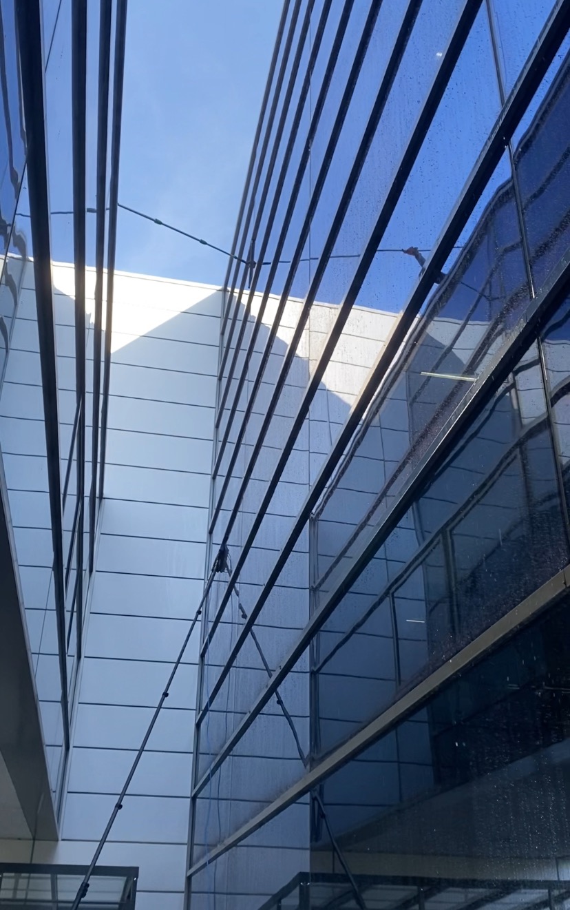 Zewnętrzne szklane ściany biurowca z widocznymi smugami wody po czyszczeniu, ujęcie z dołu na budynek z użyciem teleskopowego kija do mycia okien.