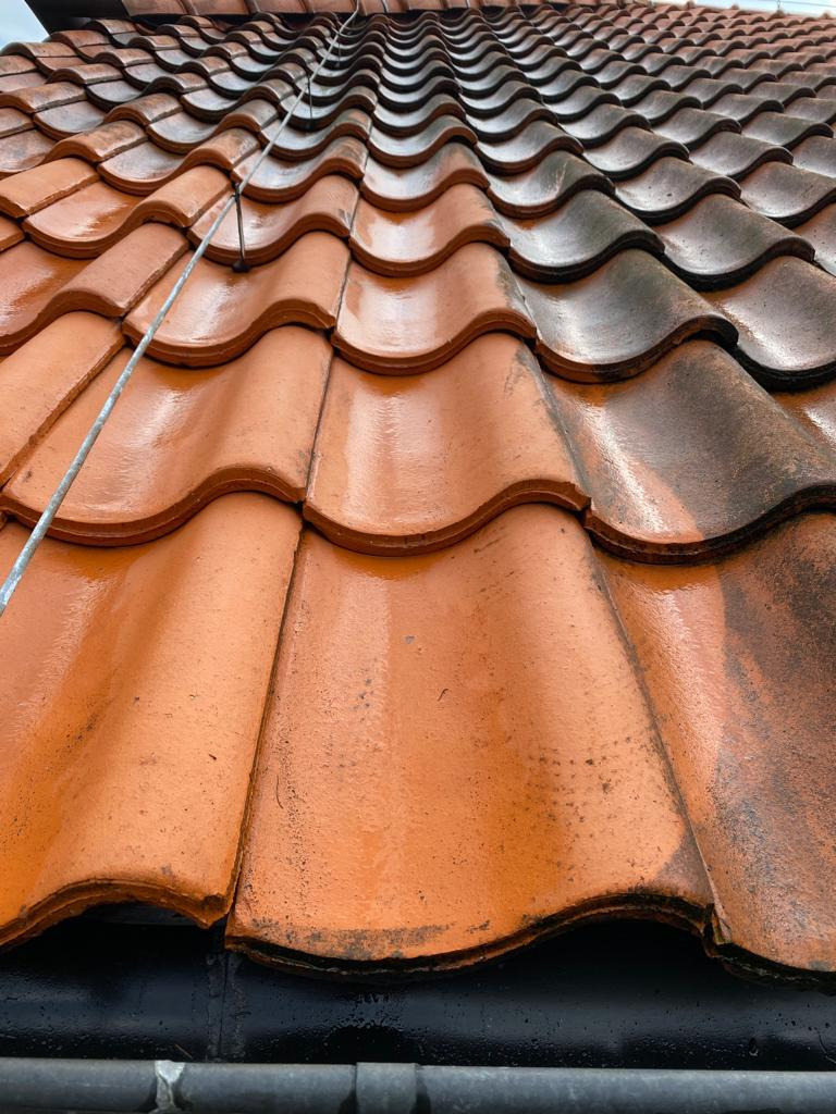 Częściowo oczyszczony dach z pomarańczowej dachówki karpiówki, widoczne różnice w kolorze pomiędzy umytą a zabrudzoną powierzchnią, z przewodem doprowadzającym wodę.