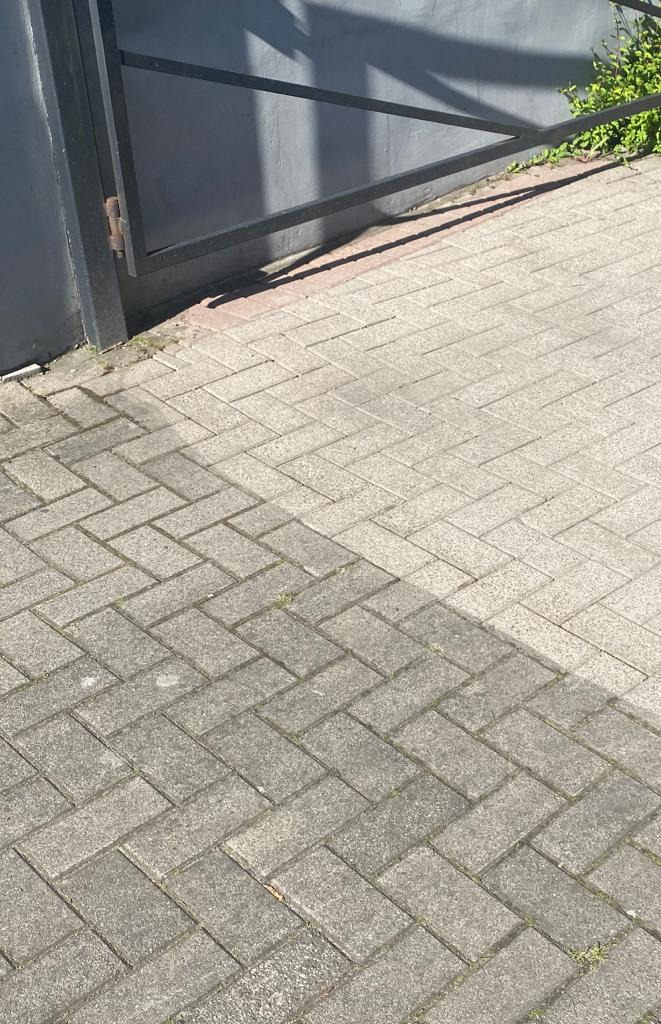 Szara kostka brukowa ułożona we wzór jodełki, częściowo zacieniona przez metalową bramę w słoneczny dzień.