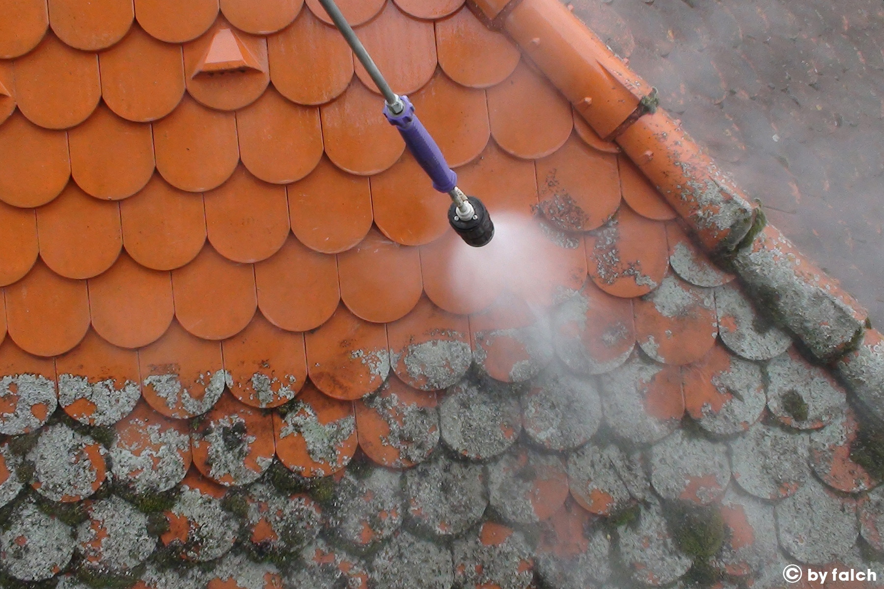 Czerwona dachówka ceramiczna częściowo pokryta mchem i porostami, czyszczona strumieniem wody pod ciśnieniem z dyszy lancy, widoczny efekt czyszczenia.