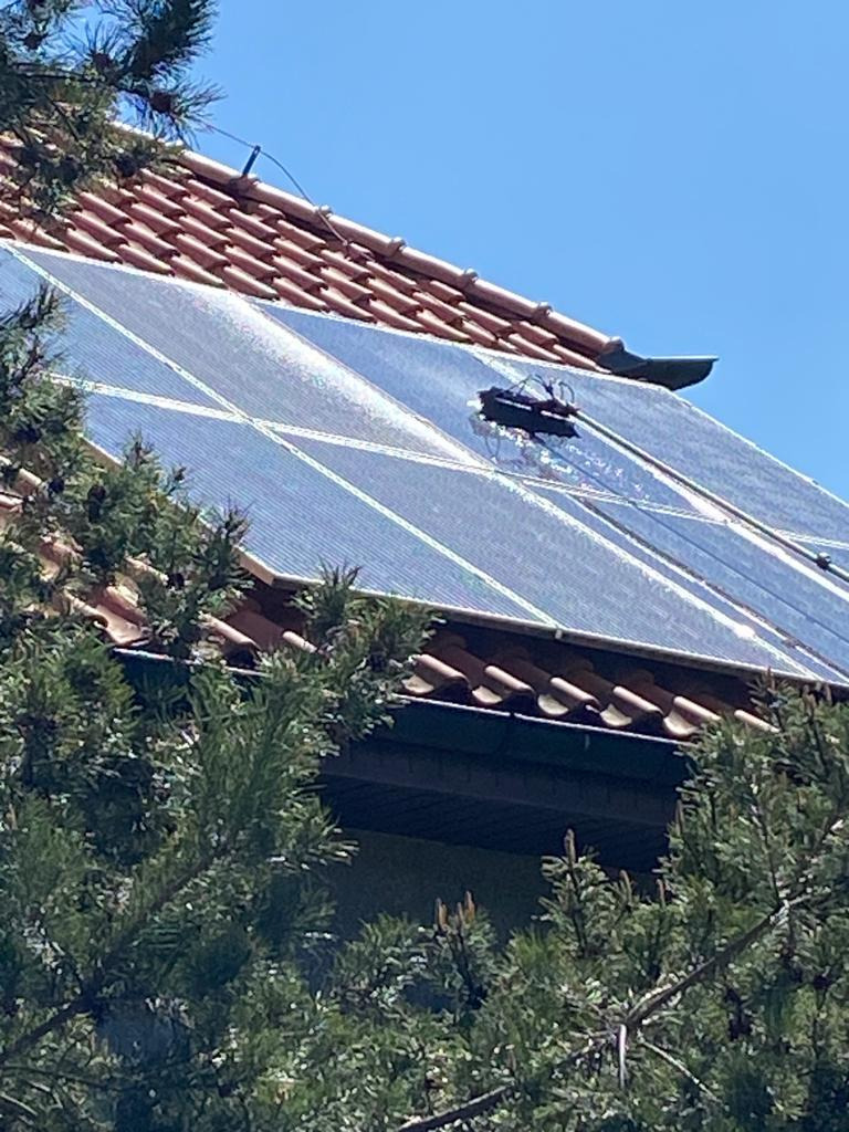 Mycie paneli słonecznych na dachu z użyciem robota czyszczącego, widok z dołu przez gałęzie drzewa.