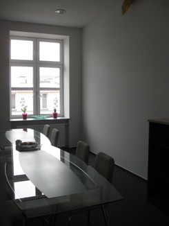 Sala konferencyjna z długim, szklanym stołem o zaokrąglonym kształcie, ustawiona w jasnym pomieszczeniu z dużym oknem. Na parapecie okiennym widoczne dwie doniczki z białymi kwiatami. Podłoga...