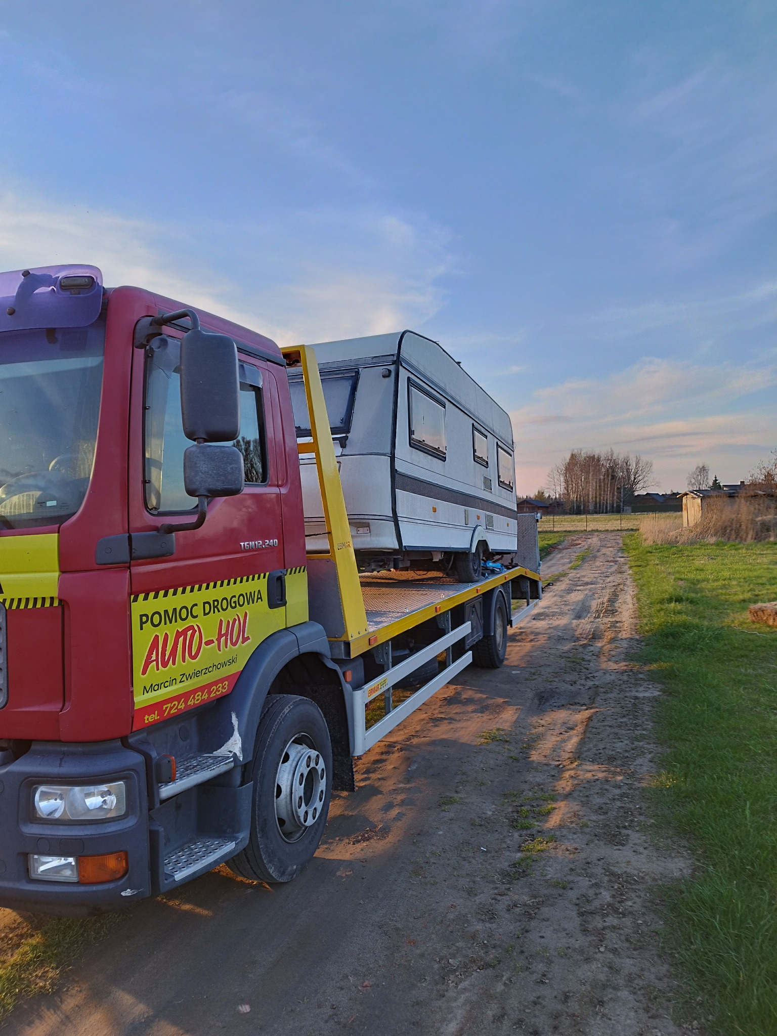 Czerwona laweta z żółtymi elementami przewozi białą przyczepę kempingową na tle polnej drogi i zachodzącego słońca.