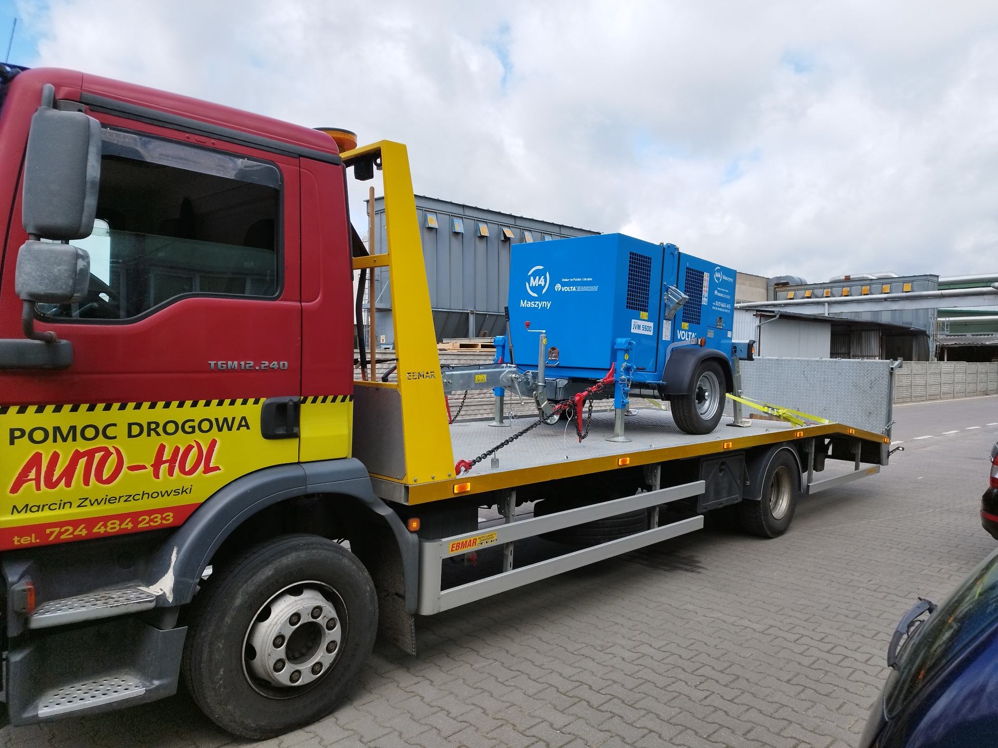 Czerwono-żółty pojazd pomocy drogowej z niebieskim agregatem prądotwórczym na platformie, zabezpieczonym łańcuchami i pasami transportowymi, na tle przemysłowego krajobrazu.