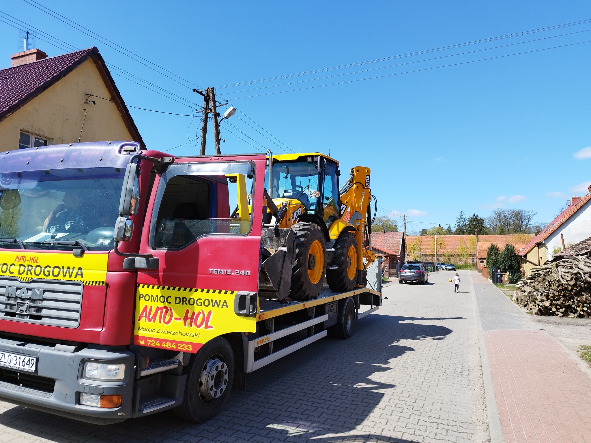 Czerwono-żółta ciężarówka pomocy drogowej z żółtą koparką JCB na platformie, zaparkowana na brukowanej ulicy w słoneczny dzień, z widocznymi domami i stertą drewna w tle.