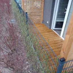 Balkon z drewnianą podłogą i ażurową osłoną, widok z góry na metalową balustradę i krzewy