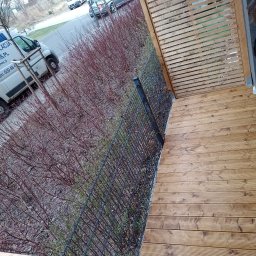 Sztuka Drewna Tworzymy z Pasją - Drewniany taras z ażurową osłoną i czarnym metalowym ogrodzeniem, widok z góry na ulicę i zaparkowane samochody.