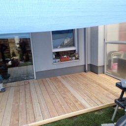 Sztuka Drewna Tworzymy z Pasją - Nowa, jasna drewniana podłoga tarasu przylegająca do domu, częściowo osłonięta niebieską plandeką, odkurzacz budowlany i stolik warsztatowy z narzędziami w tle.