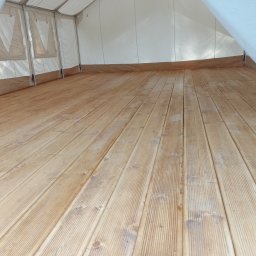 Sztuka Drewna Tworzymy z Pasją - Wnętrze namiotu z drewnianą podłogą z desek o jasnym odcieniu, widoczne ślady użytkowania i naturalne sęki w drewnie, ściany namiotu wykonane z białego, półprzezroczystego materiału.