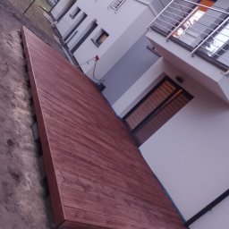 Sztuka Drewna Tworzymy z Pasją - Nowo wybudowany taras z desek w odcieniach czerwieni przylegający do nowoczesnego domu z balkonem z metalową balustradą, widoczne przygotowane podłoże.