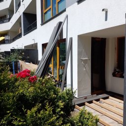 Sztuka Drewna Tworzymy z Pasją - W budowie taras przy nowoczesnym apartamentowcu: drewniane legary ułożone na betonowej powierzchni, obok leżą panele kompozytowe, poziomica i worek ze śrubami.